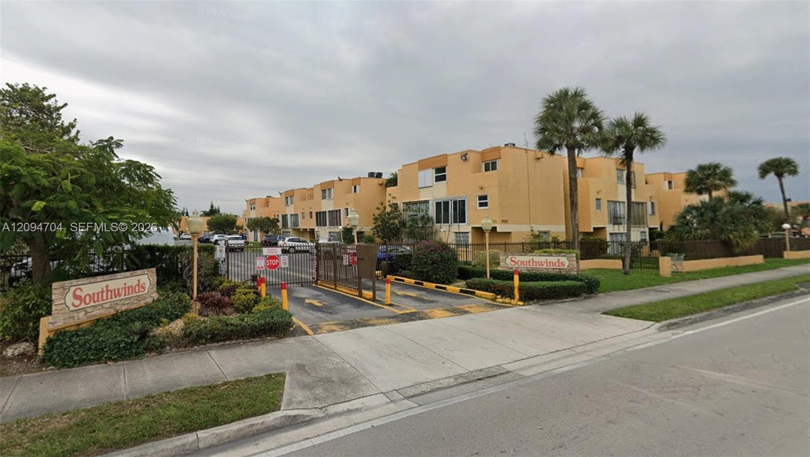 Gated entrance featuring a metal security gate, concrete driveway, stone-veneer signage, and manicured landscaping with palm trees