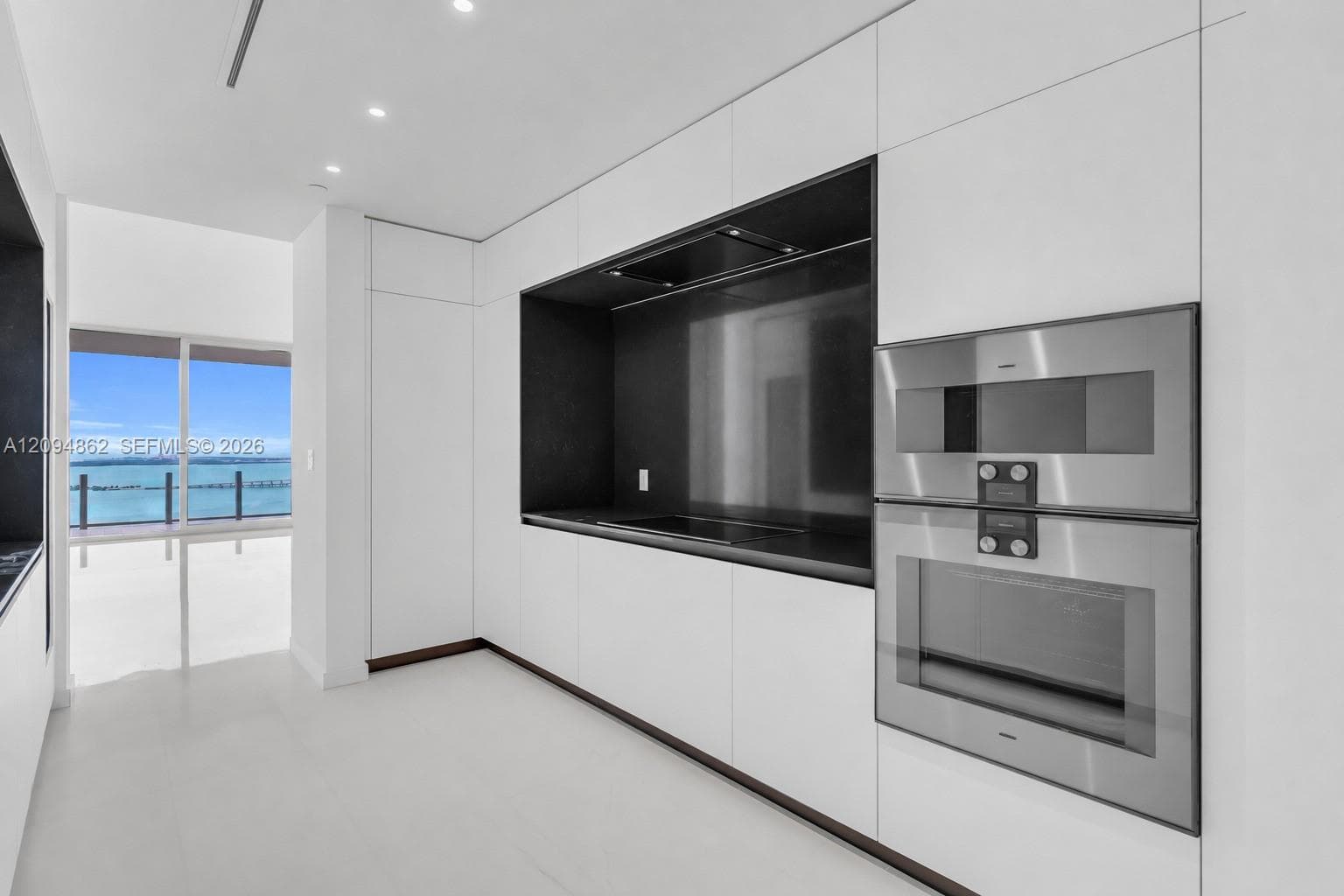 Minimalist kitchen featuring matte white flat-panel cabinetry, integrated stainless steel double wall ovens, a contrasting dark cooktop backsplash, and polished tile flooring