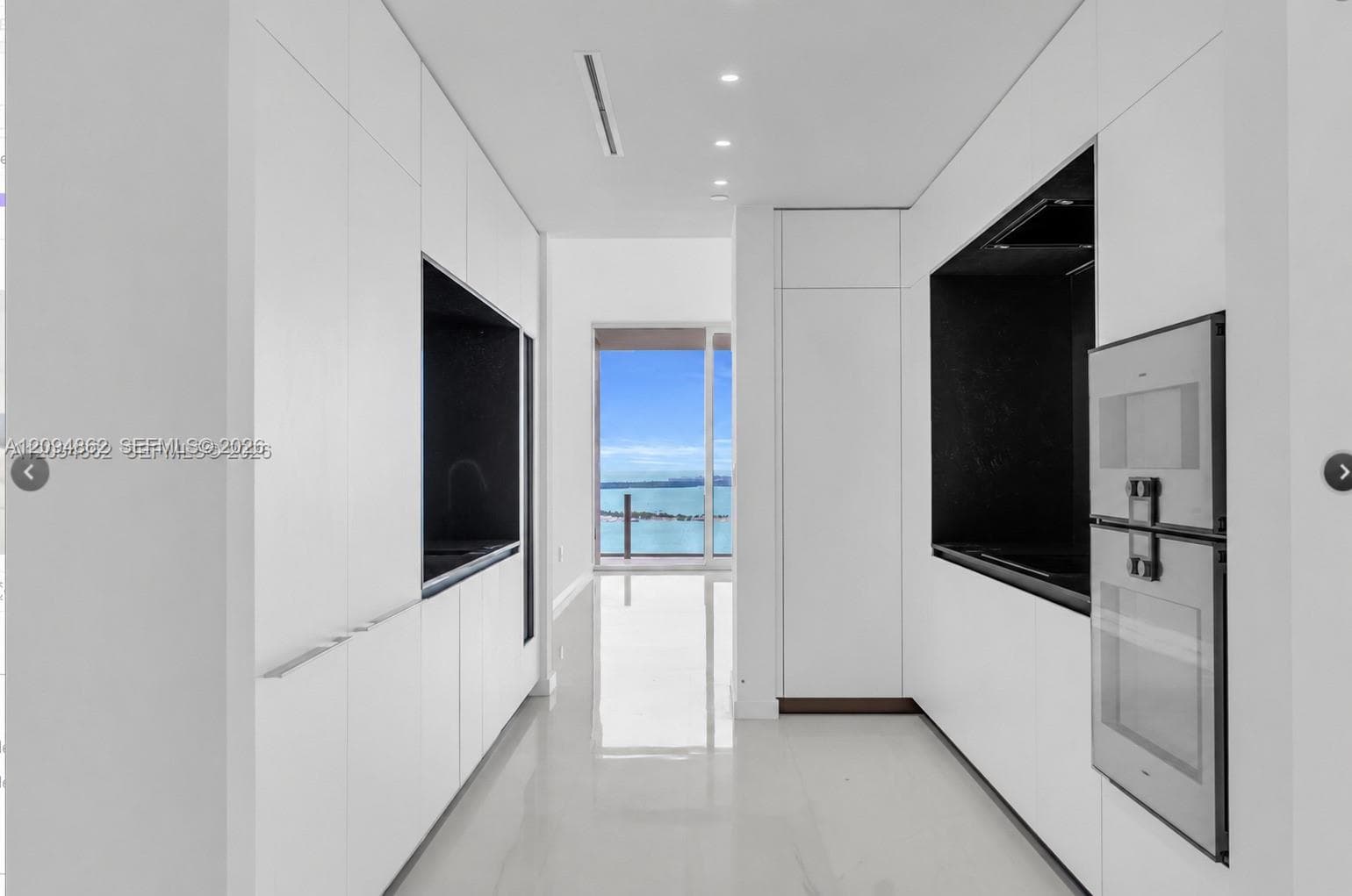 Sleek galley kitchen featuring flat-panel white cabinetry, black contrast alcoves, integrated stainless steel appliances, recessed ceiling lighting, and polished stone flooring