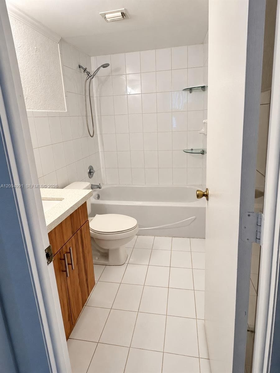 Bathroom featuring a wood-finish vanity with a light-toned countertop, white tile flooring, white wall tiling, and a built-in bathtub with a shower head and glass shelving