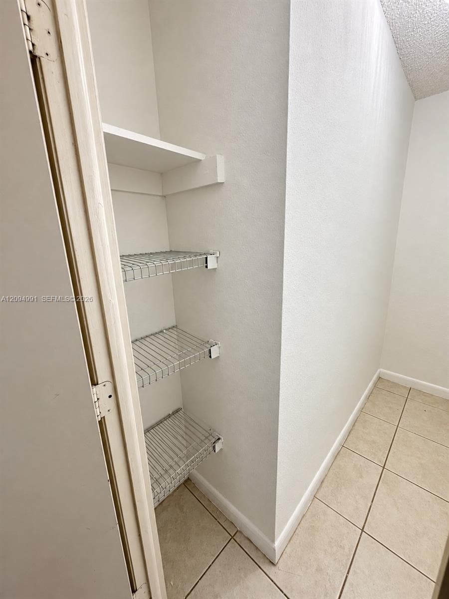 Storage area featuring neutral tile flooring and white walls, equipped with three levels of white wire shelving and one fixed shelf