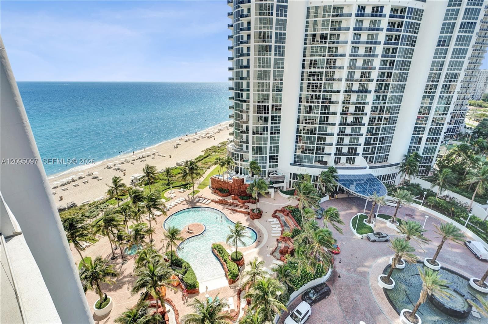 High-rise architectural exterior featuring glass-paned facades, a landscaped courtyard with a curvilinear swimming pool, mature palm trees, a circular driveway, and direct oceanfront positioning