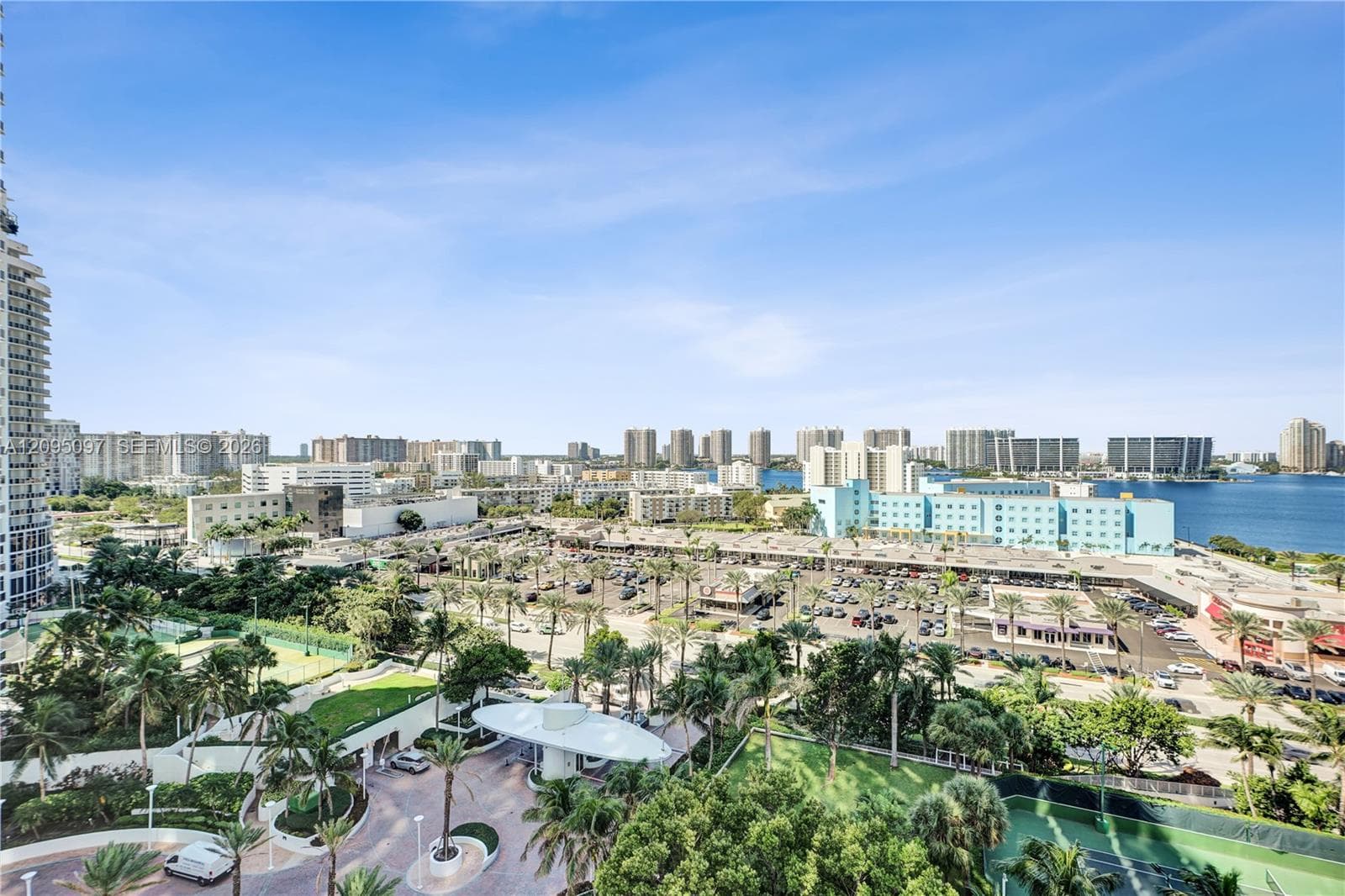 Elevated exterior perspective featuring a circular porte-cochère, paved motor court, mature landscaping, and tennis courts