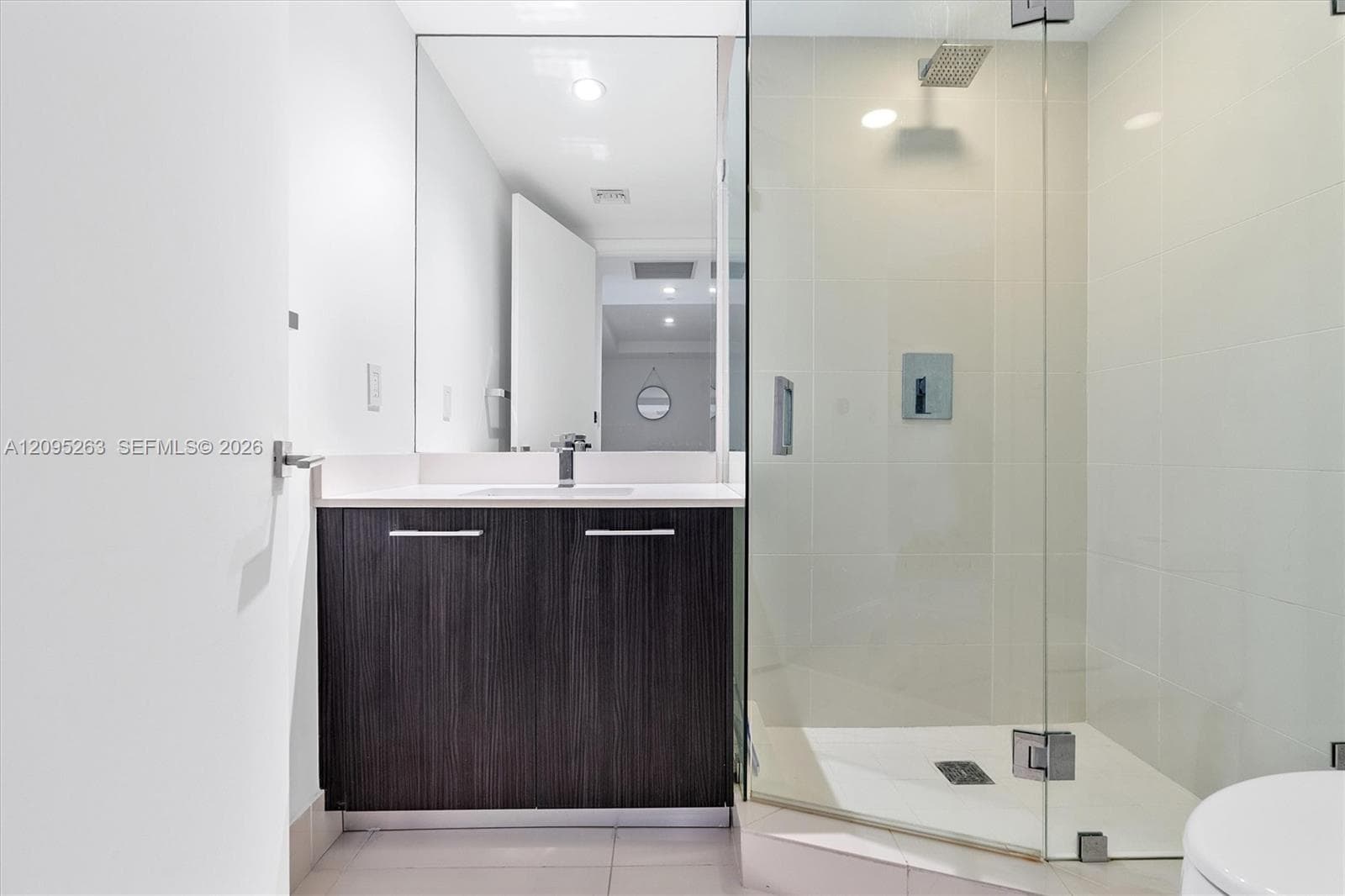 Dark wood-finish vanity with a light-toned countertop, large-format mirror, glass-enclosed shower, and wall-mounted rainfall shower head