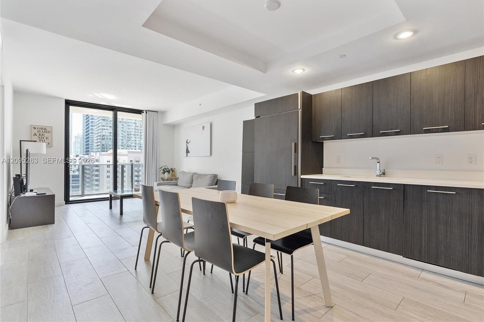 Open-concept living space featuring wood-finish floor tiling, recessed lighting, dark flat-panel cabinetry, integrated refrigerator, and a glass-paneled sliding door leading to an exterior balcony