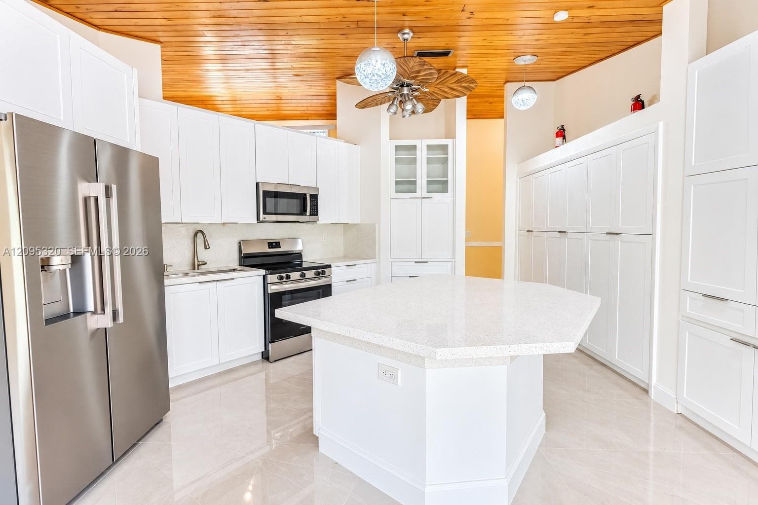 Wood-panel ceiling with a ceiling fan, white cabinetry with stainless steel appliances, and a central island with a light-colored countertop, complemented by light-toned flooring