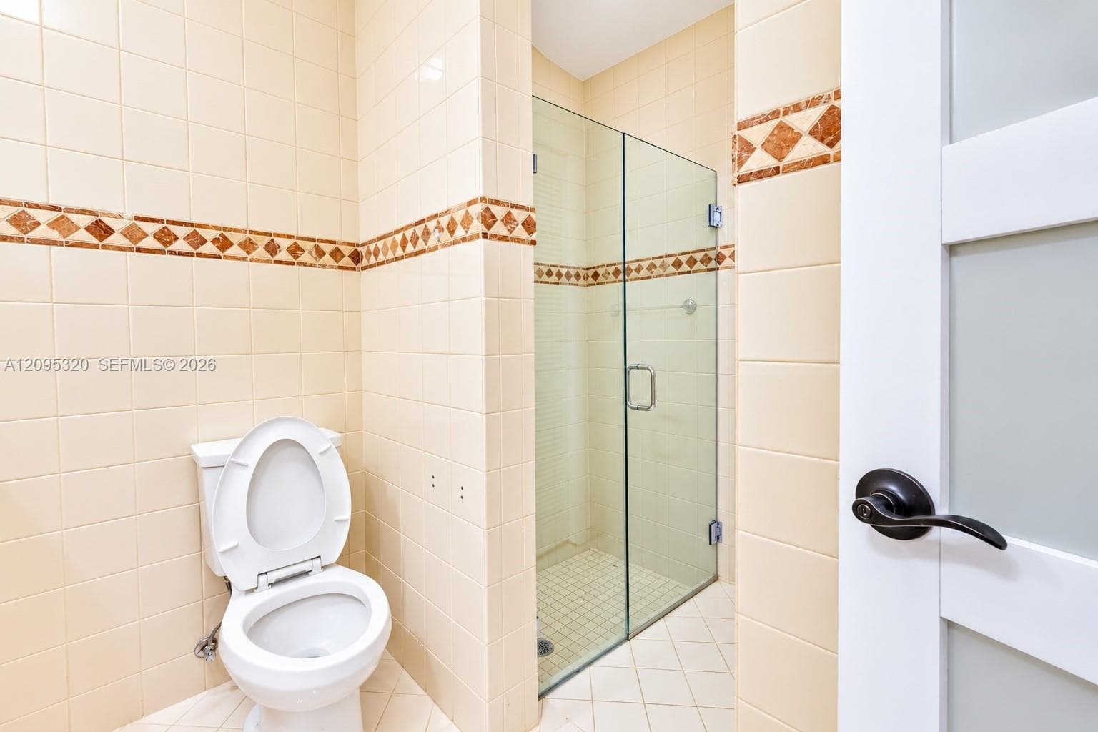 Tiled bathroom featuring a glass-enclosed shower, decorative tile border, white toilet, and white door with dark hardware