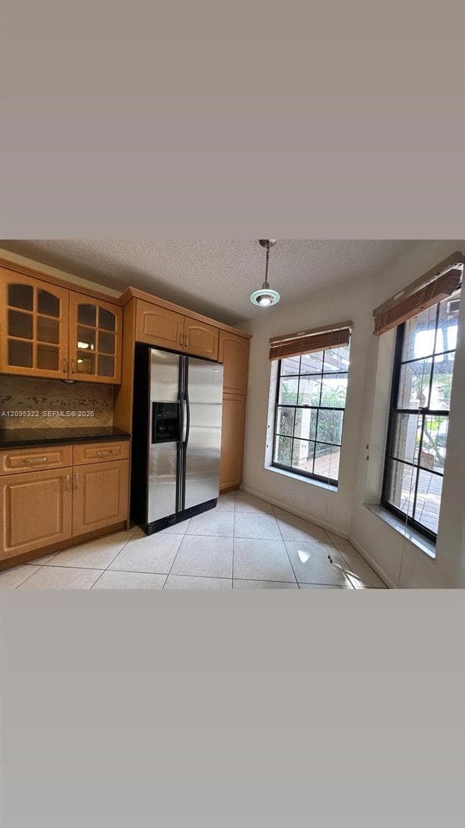 Kitchen featuring wood cabinetry with glass-panel upper doors, stone-finish countertops, stainless steel side-by-side refrigerator, tile flooring, and windows with integrated grilles