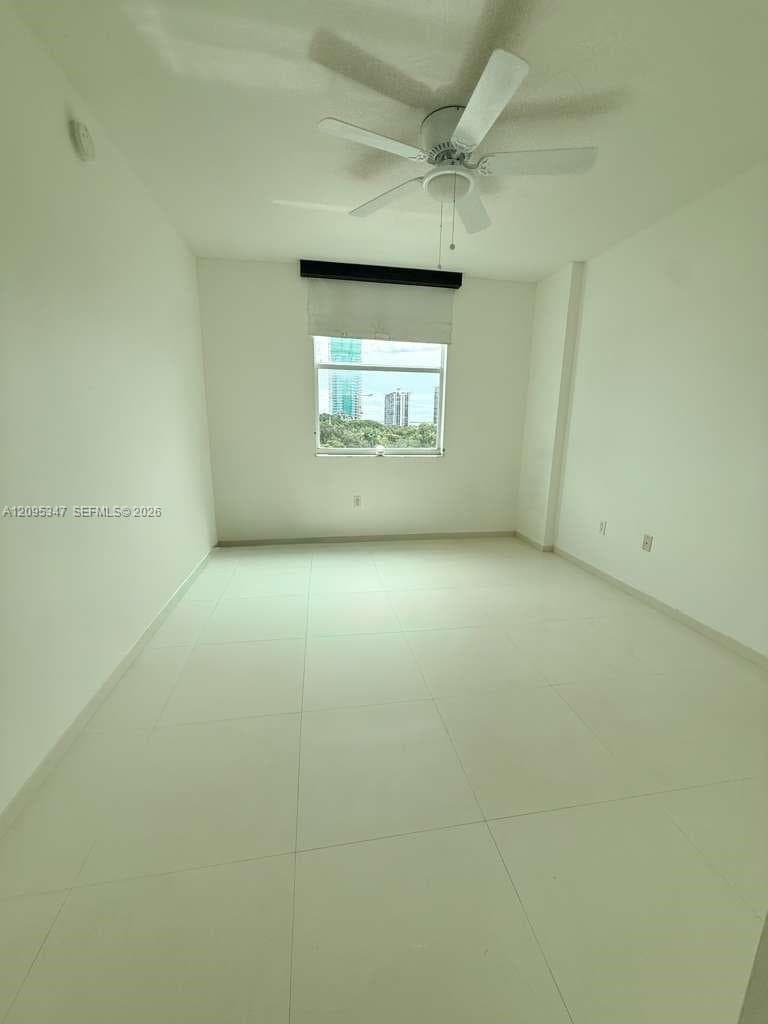 Neutral-toned room featuring large-format tile flooring, a white ceiling fan, and a single window with a roller shade