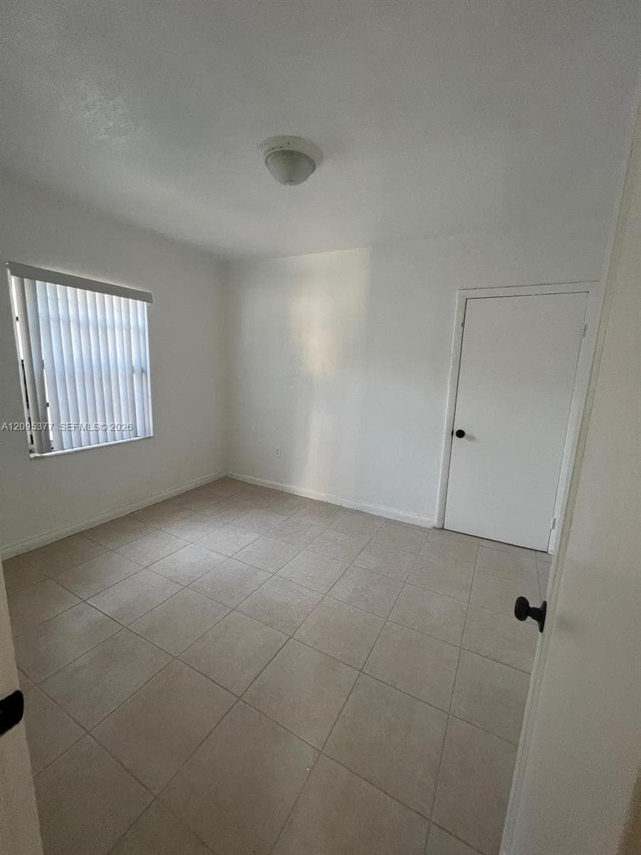 Neutral-toned tile flooring, white wall finish, interior door with black hardware, and a single window featuring vertical blinds