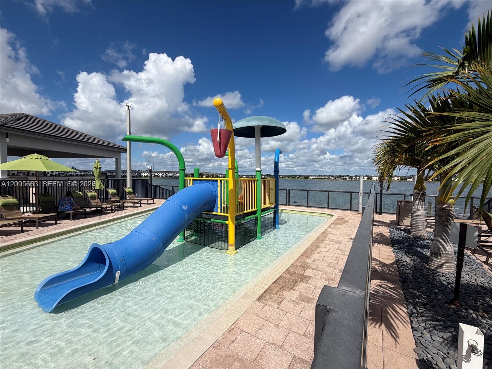Waterside splash pad featuring a blue tube slide, overhead dumping bucket, and mushroom water feature, finished with paved decking and perimeter metal fencing