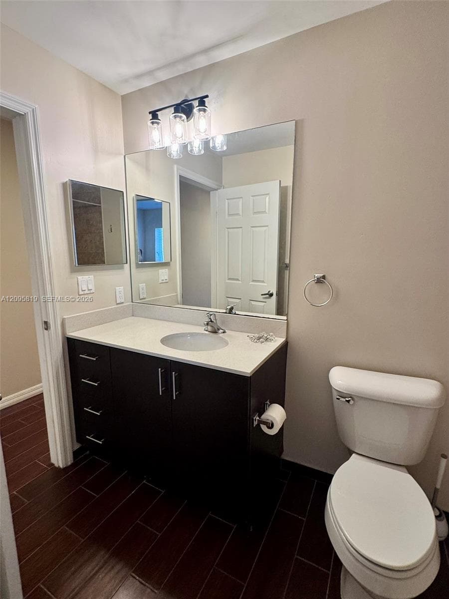 Dark wood-finish cabinetry topped with a solid surface counter, integrated sink, large wall-mounted mirror, and wood-finish tile flooring
