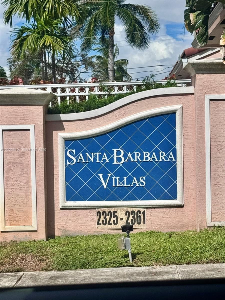 Stucco-finished masonry entrance monument featuring a custom blue tile inlay with white decorative grid pattern, integrated pillar caps, and established tropical landscaping