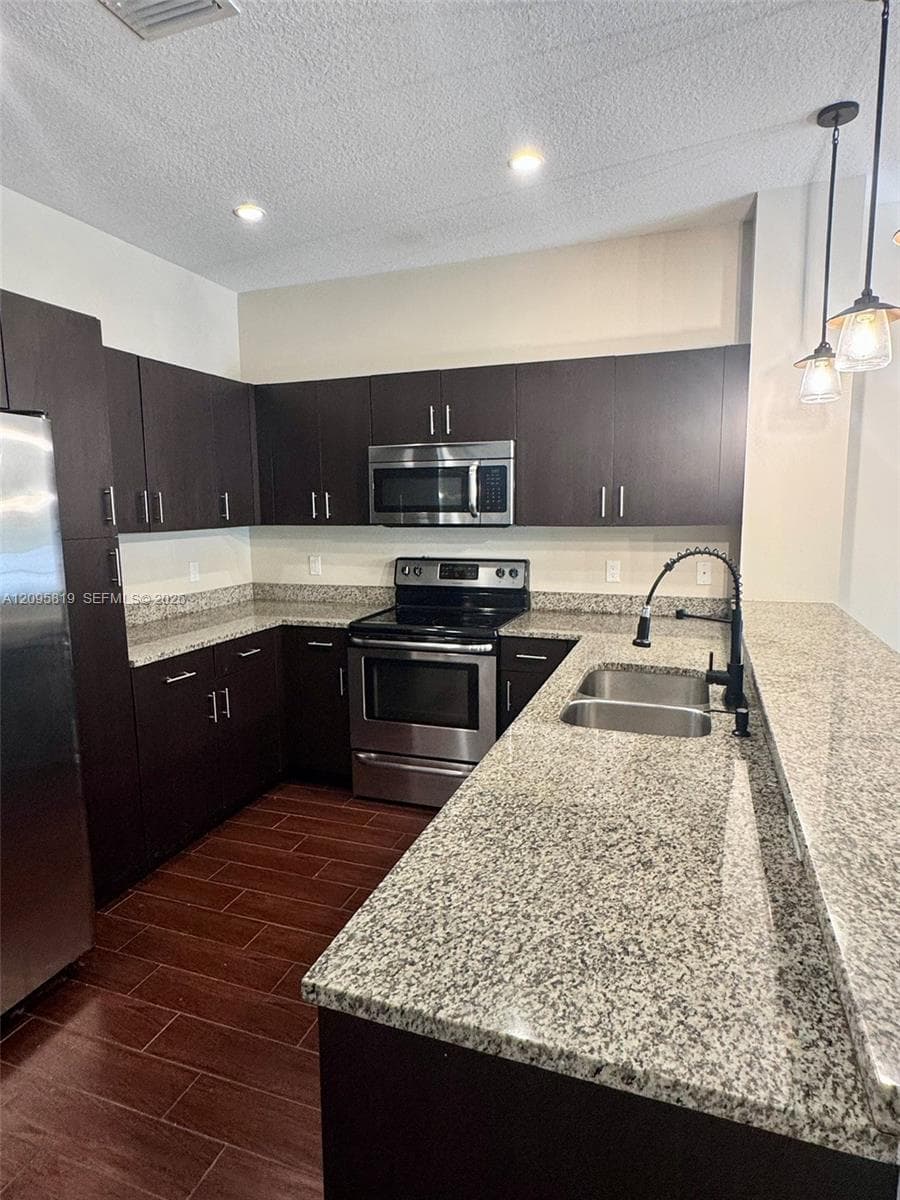 Kitchen featuring dark cabinetry, stone-finish countertops, stainless steel appliances, wood-finish tile flooring, and pendant lighting over a dual-basin sink