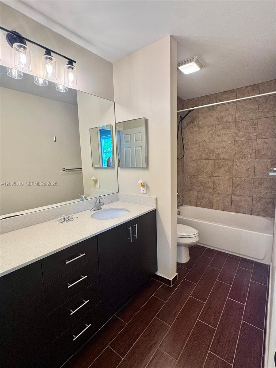 Modern vanity with a light-toned countertop, dark cabinetry, wood-finish flooring, a full-width mirror with integrated lighting, and a tile-surround bathtub