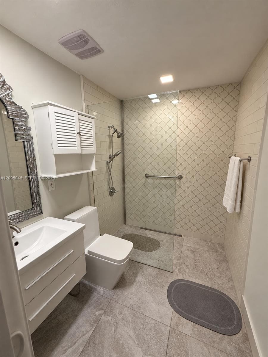 Modern white vanity with a rectangular sink, wall-mounted cabinet, walk-in shower with a glass panel, patterned tile accent wall, and large-format tile flooring