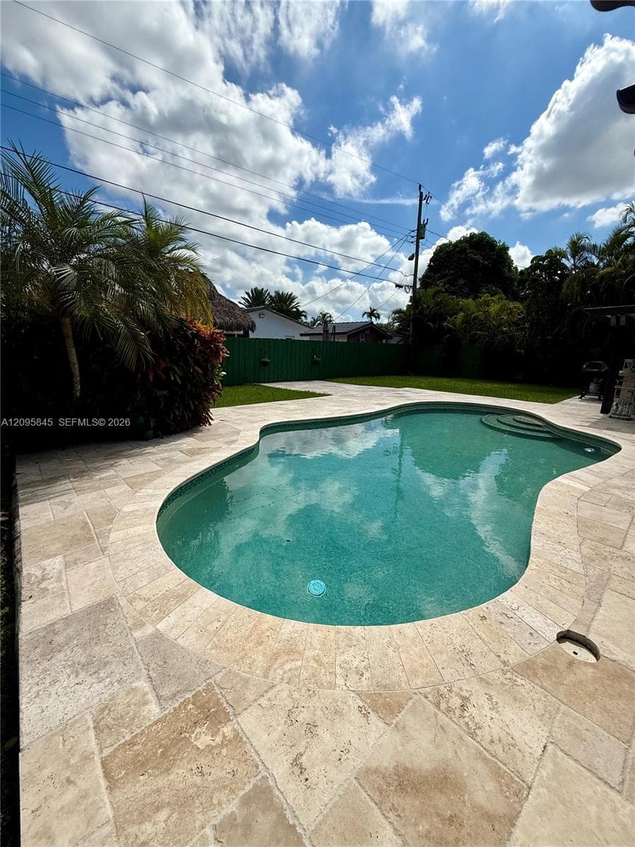 In-ground freeform swimming pool with stone paver decking, green privacy fencing, and mature landscaping