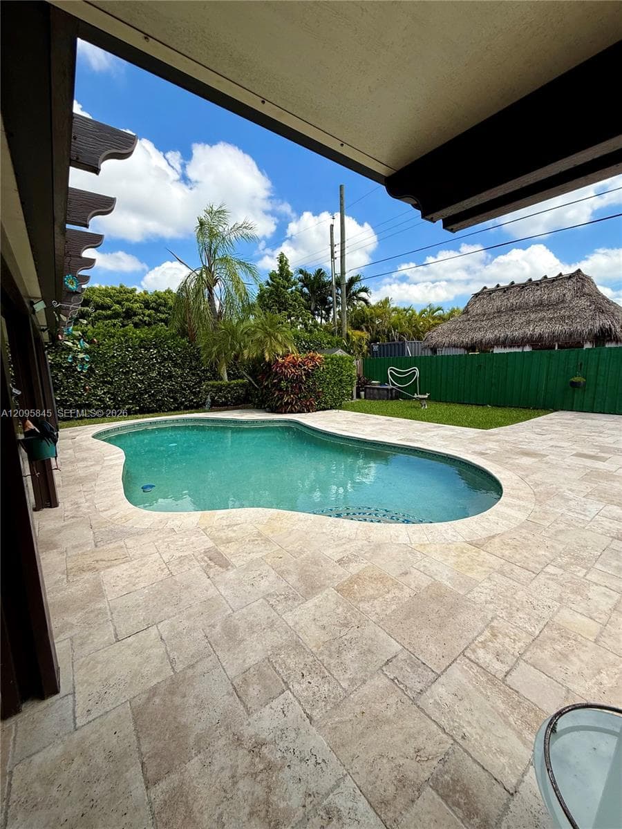 Custom-shaped swimming pool with stone paver decking, covered patio, privacy hedging, and detached thatched-roof structure