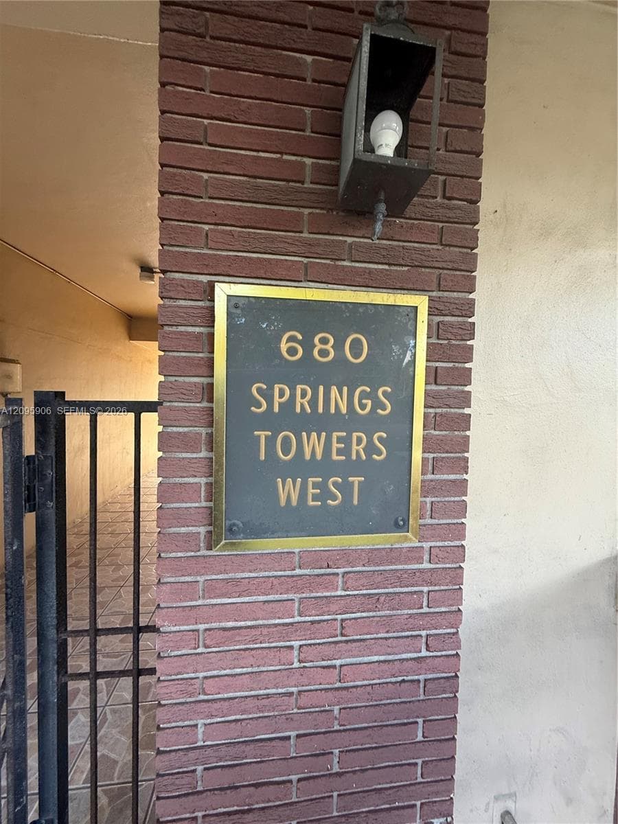 Exterior brick pillar entrance featuring a metallic building identification plaque, black metal gate, and attached wall-mounted lantern