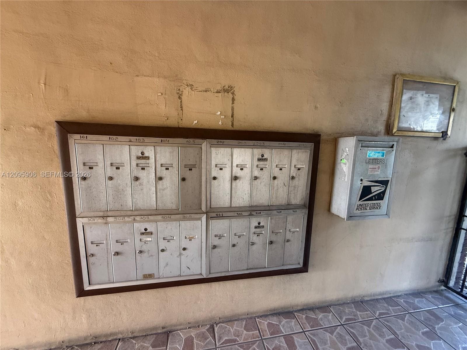 Wall-mounted bank of twenty metal mailboxes with a dedicated outgoing mail collection unit and a framed notice board