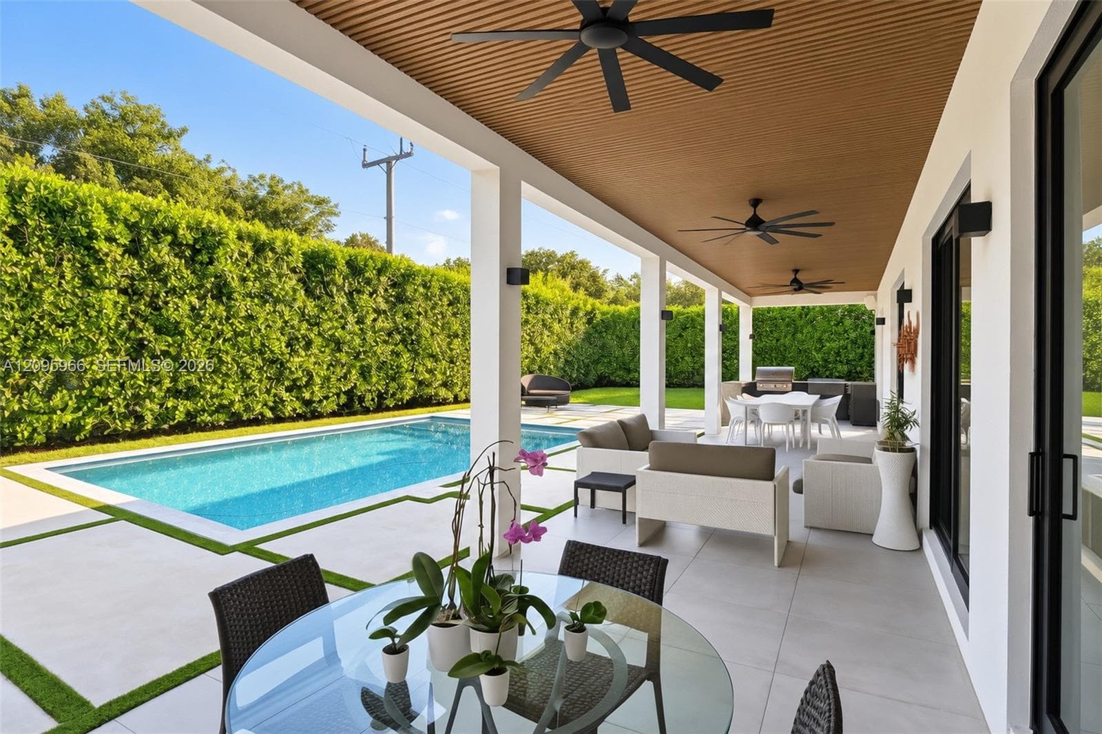 Covered patio featuring a wood-slat ceiling, recessed ceiling fans, large-format light tile flooring, an integrated outdoor kitchen, and a swimming pool surrounded by paved decking with turf inlays