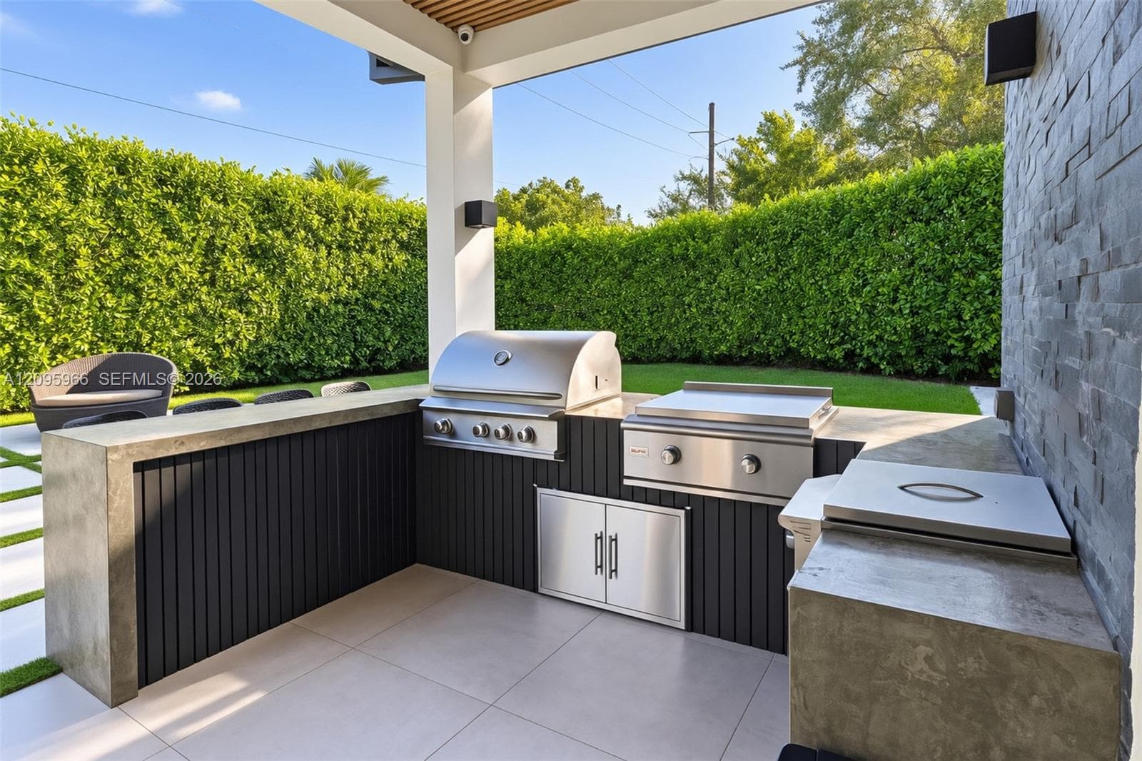 Covered outdoor kitchen featuring a built-in stainless steel grill, a flat-top griddle, concrete countertops, vertical slat cabinetry, and a stacked stone accent wall