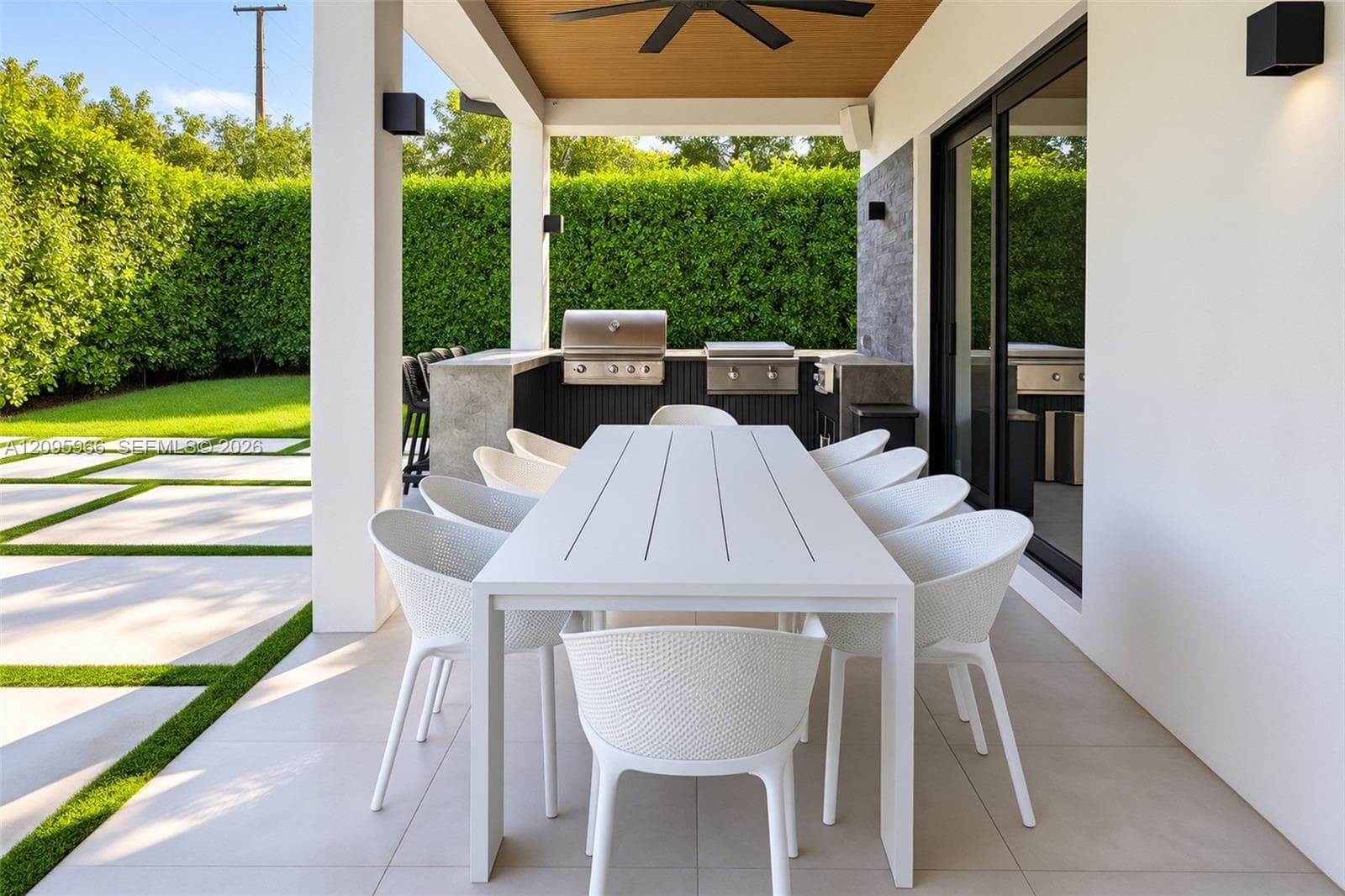 Covered patio featuring large-format tile flooring, a wood-paneled ceiling with an integrated fan, built-in outdoor kitchen with stainless steel appliances, and a concrete patio surface with turf accents