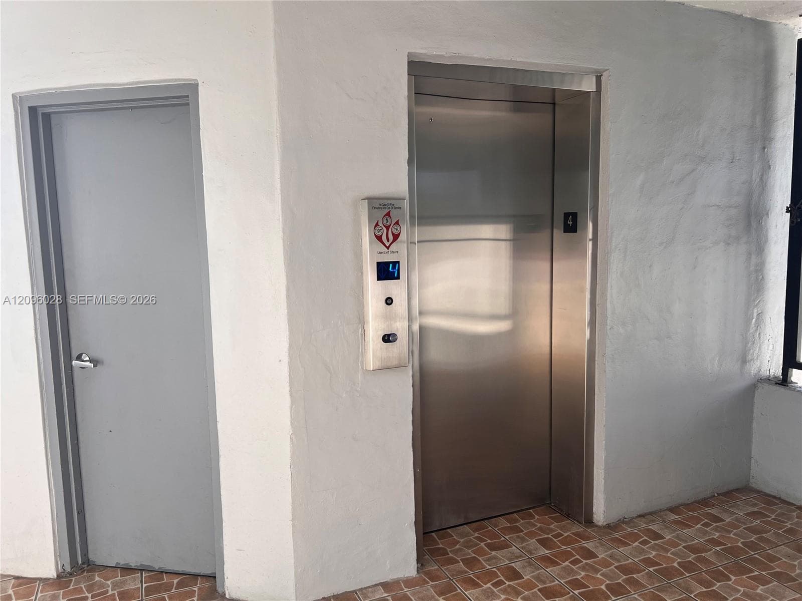 Stainless steel elevator with a digital floor indicator and push-button controls, accompanied by a solid-core door and terracotta-tone tiled flooring
