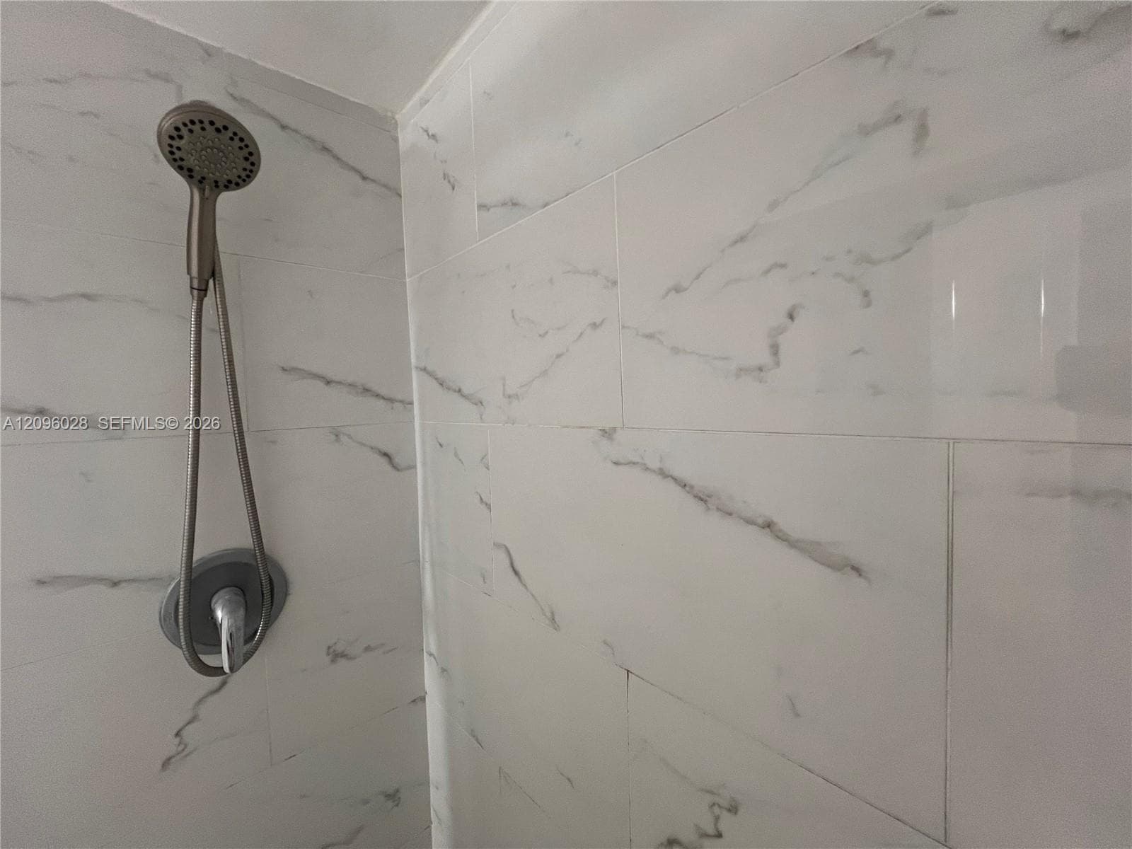 Shower surround featuring large-format, light-toned stone-finish tile with integrated veining, complemented by a metallic handheld showerhead and circular wall-mounted control valve