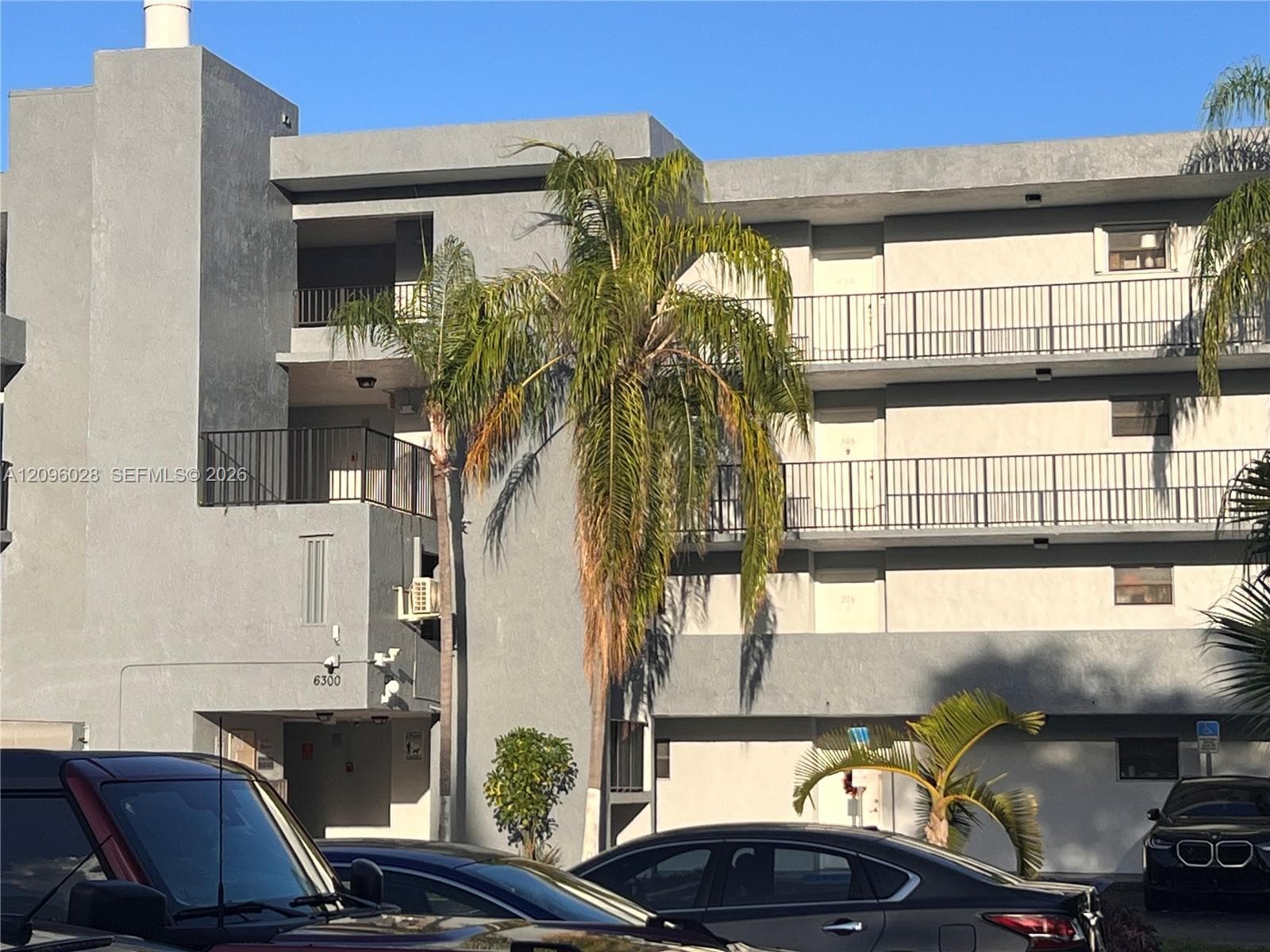 Multi-story exterior featuring a gray stucco facade, private balconies with black metal railings, and mature palm trees
