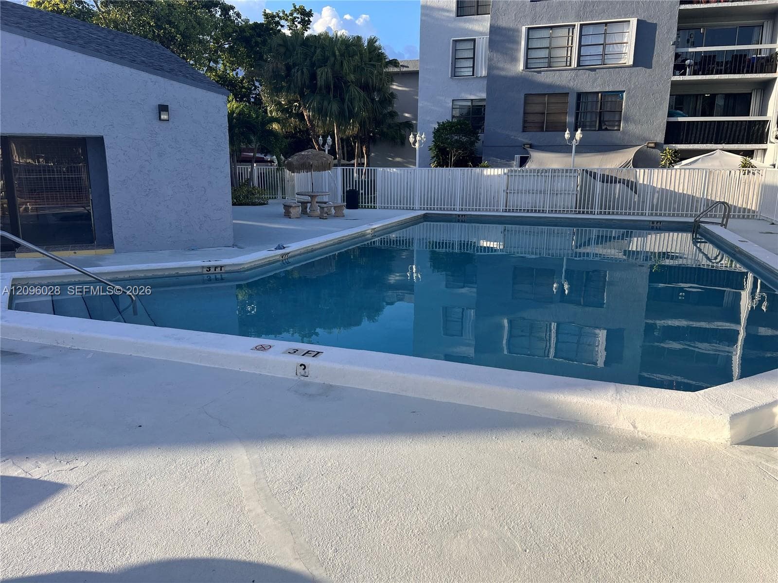In-ground swimming pool featuring a multi-sided basin, metal entry handrails, and a wide concrete deck enclosed by white fencing