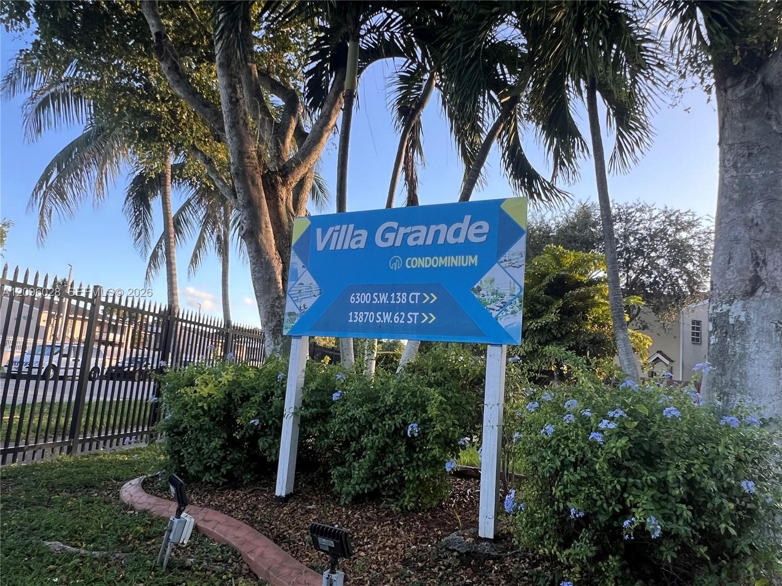 Exterior signage for Villa Grande Condominium featuring mature palm trees, perimeter metal fencing, and landscaped shrubbery
