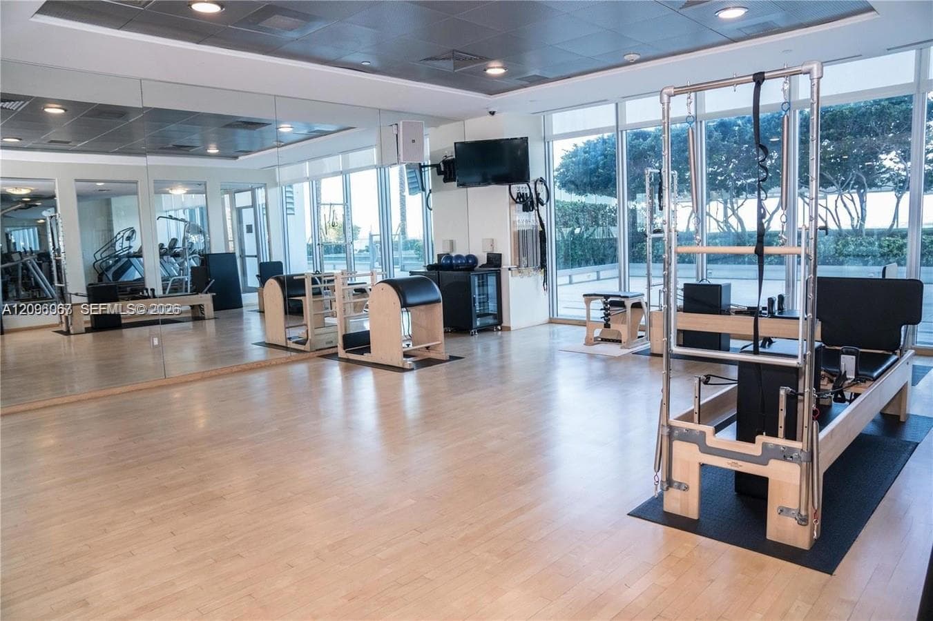 Expansive fitness studio featuring gleaming wood-finish flooring, floor-to-ceiling windows with natural light, modern ceiling-mounted lighting, and specialized Pilates equipment stations