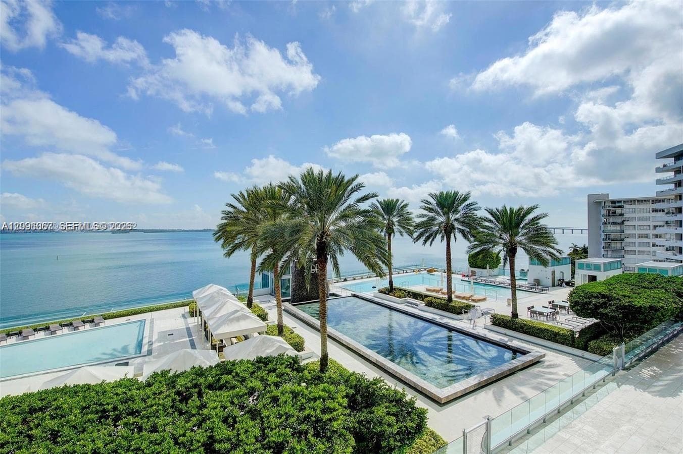 Expansive waterfront hardscaping featuring two swimming pools, mature palm trees, and shaded cabana areas