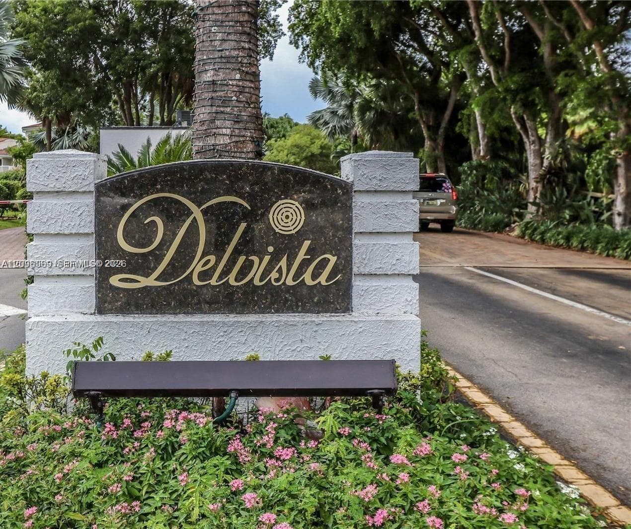 Community signage featuring a dark stone plaque with gold lettering, set within a textured white masonry monument base, surrounded by manicured landscaping and mature trees