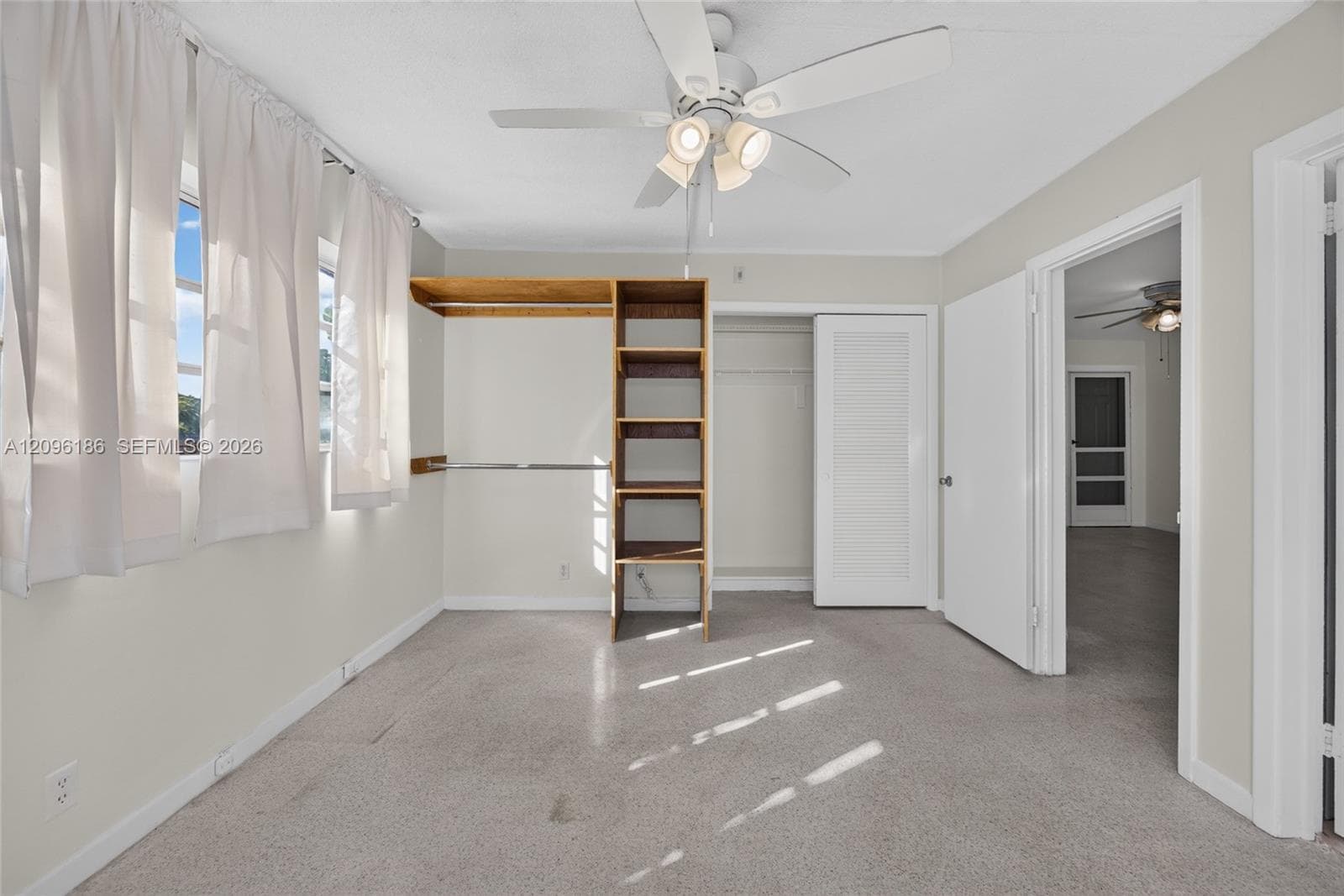 Neutral-toned room featuring a central ceiling fan with integrated lighting, open built-in shelving, carpeted flooring, and a closet with a louvered door