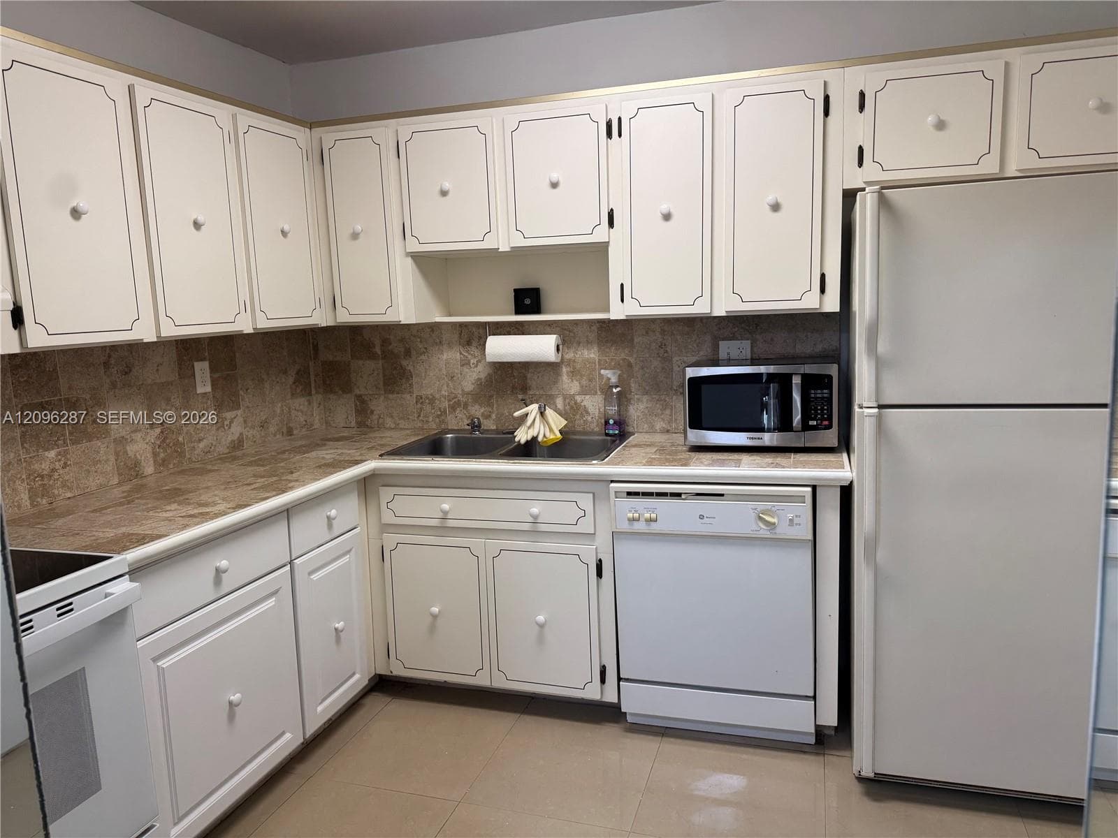 Kitchen featuring white cabinetry with decorative trim, neutral-toned stone-finish tile countertops, a coordinating stone tile backsplash, a stainless steel double-basin sink, and large-format tile flooring