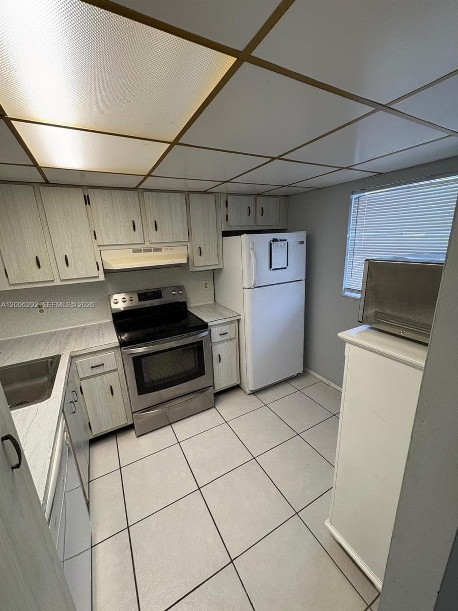 Kitchen featuring light-finish cabinetry, stainless steel range, white tile flooring, drop ceiling, and surface-mount lighting