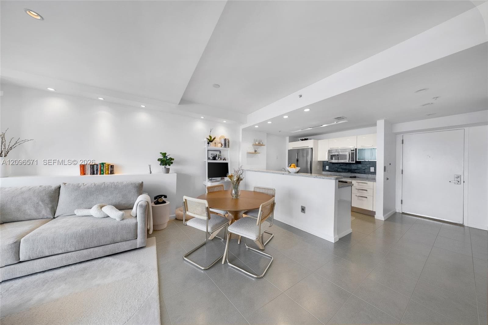 Open-concept interior featuring large-format tile flooring, recessed ceiling lighting, a kitchen peninsula with a stone countertop, white flat-panel cabinetry, and a built-in display niche