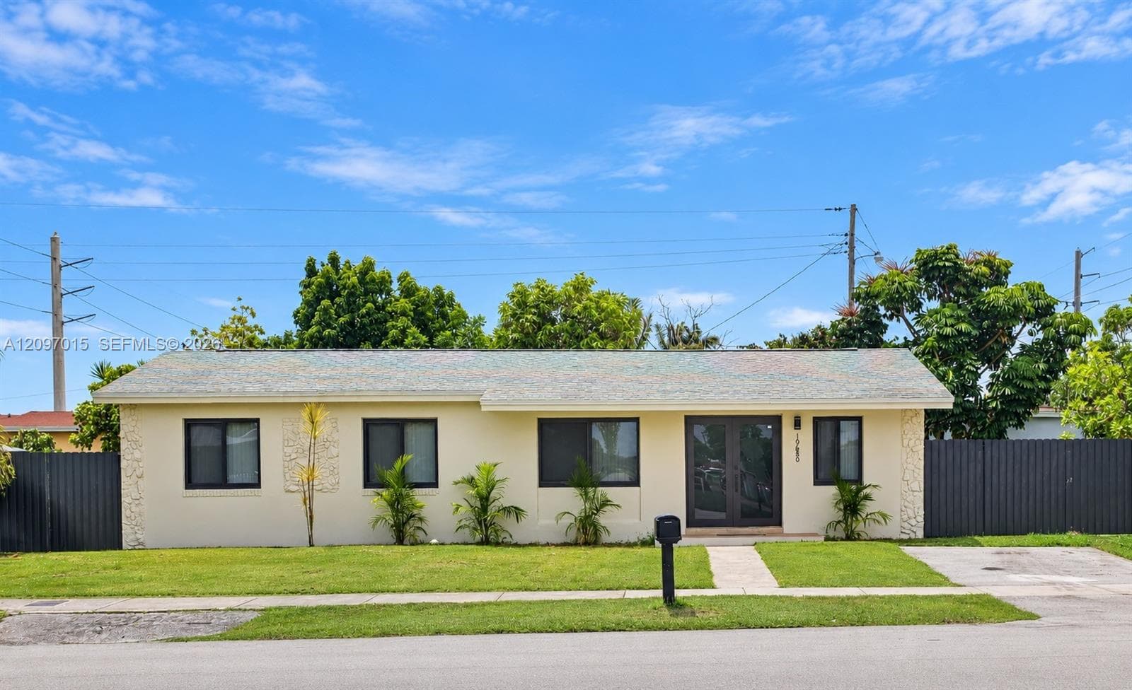 Single-story residence featuring a light-colored stucco exterior, stone accent panels, dark-framed windows, and double-entry doors, enclosed by perimeter fencing