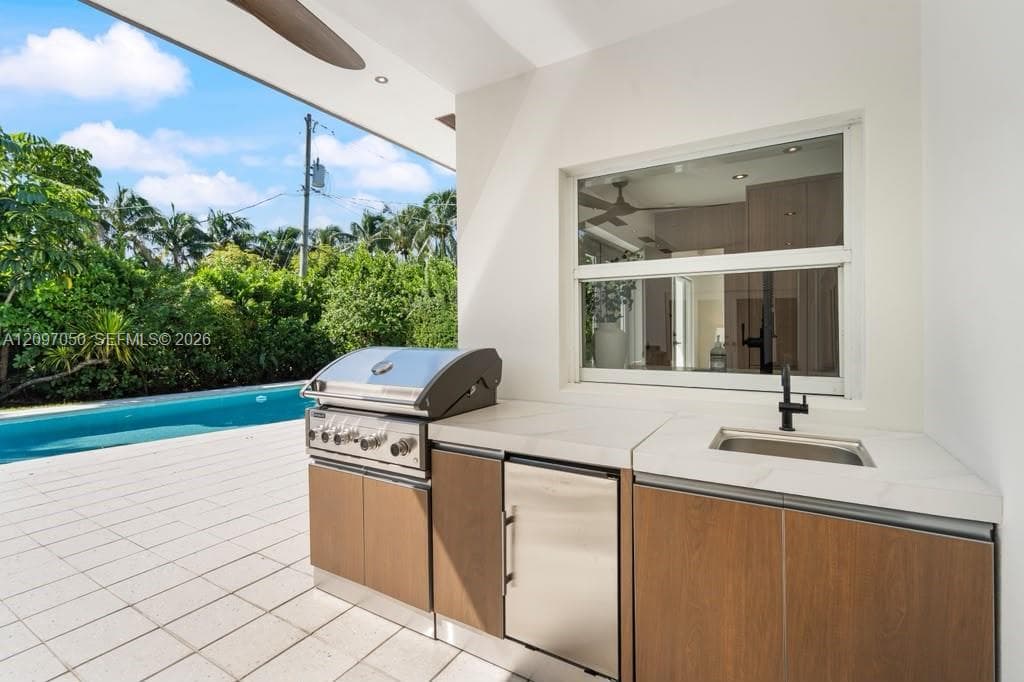 Outdoor kitchen featuring a built-in stainless steel grill, stone countertop, undermount sink with matte black faucet, and wood-finish cabinetry