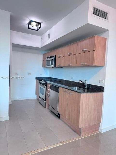Kitchen featuring wood-finish cabinetry, dark stone countertops, stainless steel appliances, tile flooring, and a box-style ceiling light fixture
