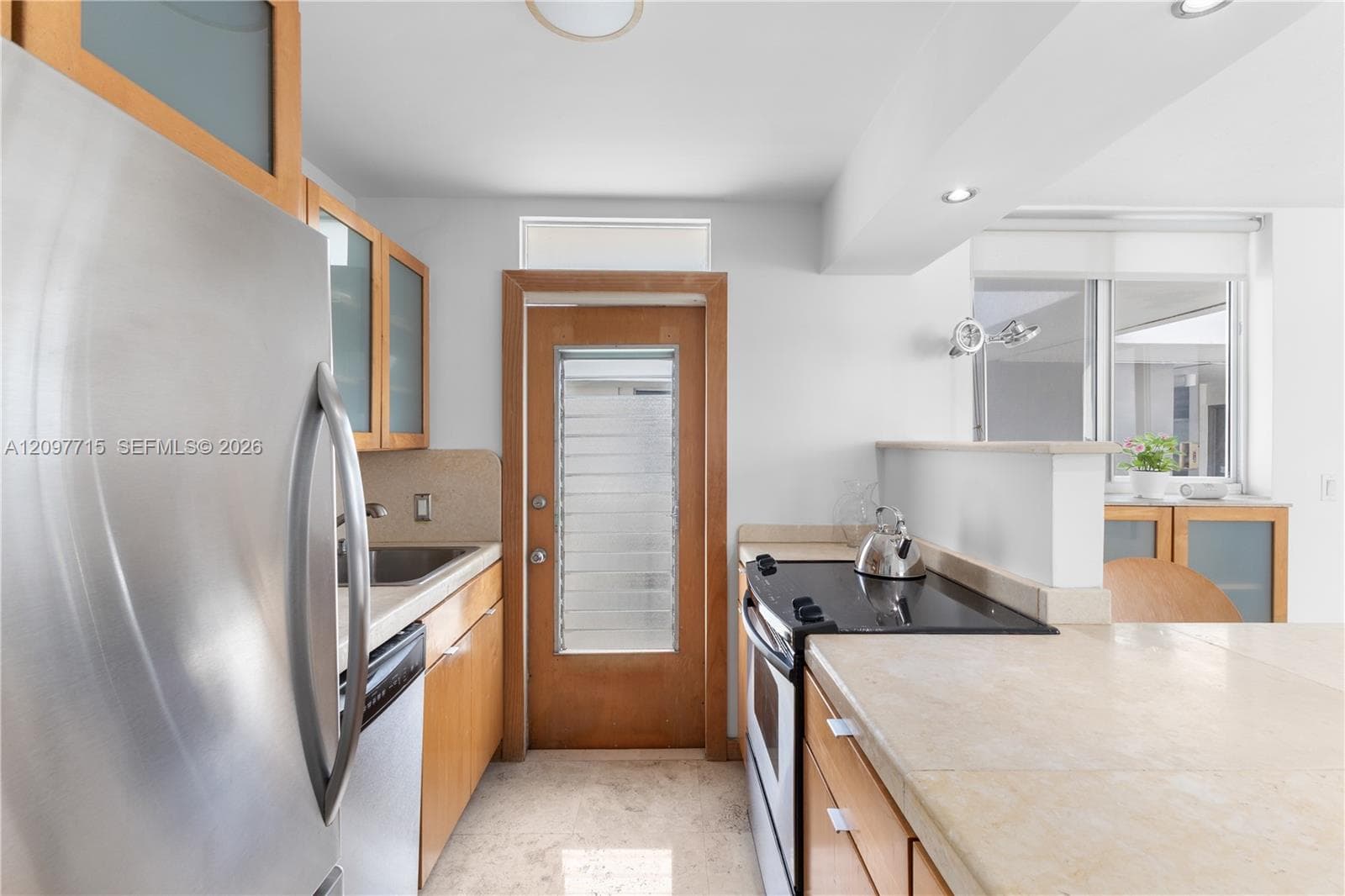 Stainless steel refrigerator, integrated cooktop, light wood-finish cabinetry, light-toned countertops, and a glass-paneled door