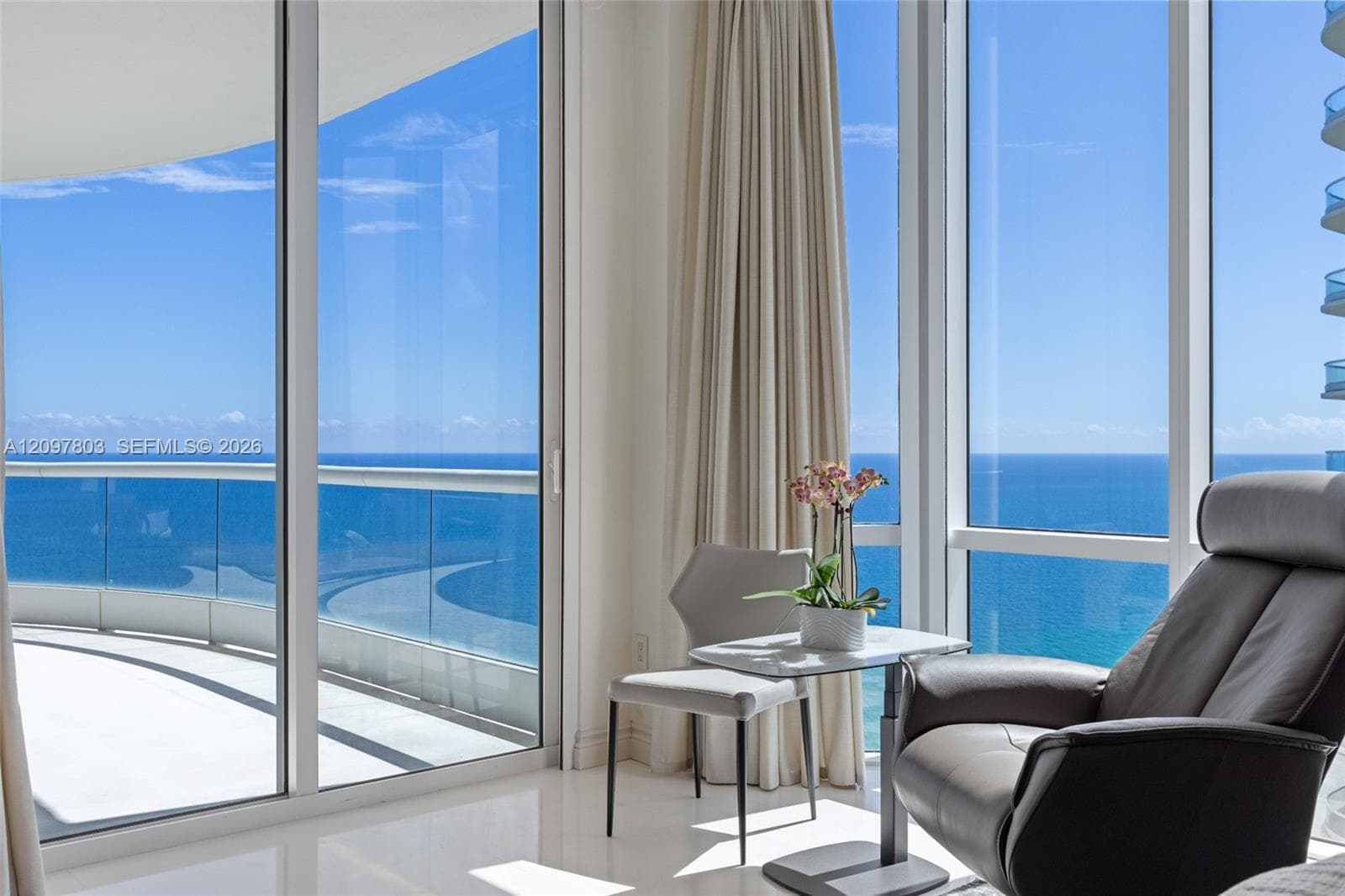 Floor-to-ceiling glass windows, sliding glass doors, and a wrap-around balcony with glass panel railing overlooking the ocean