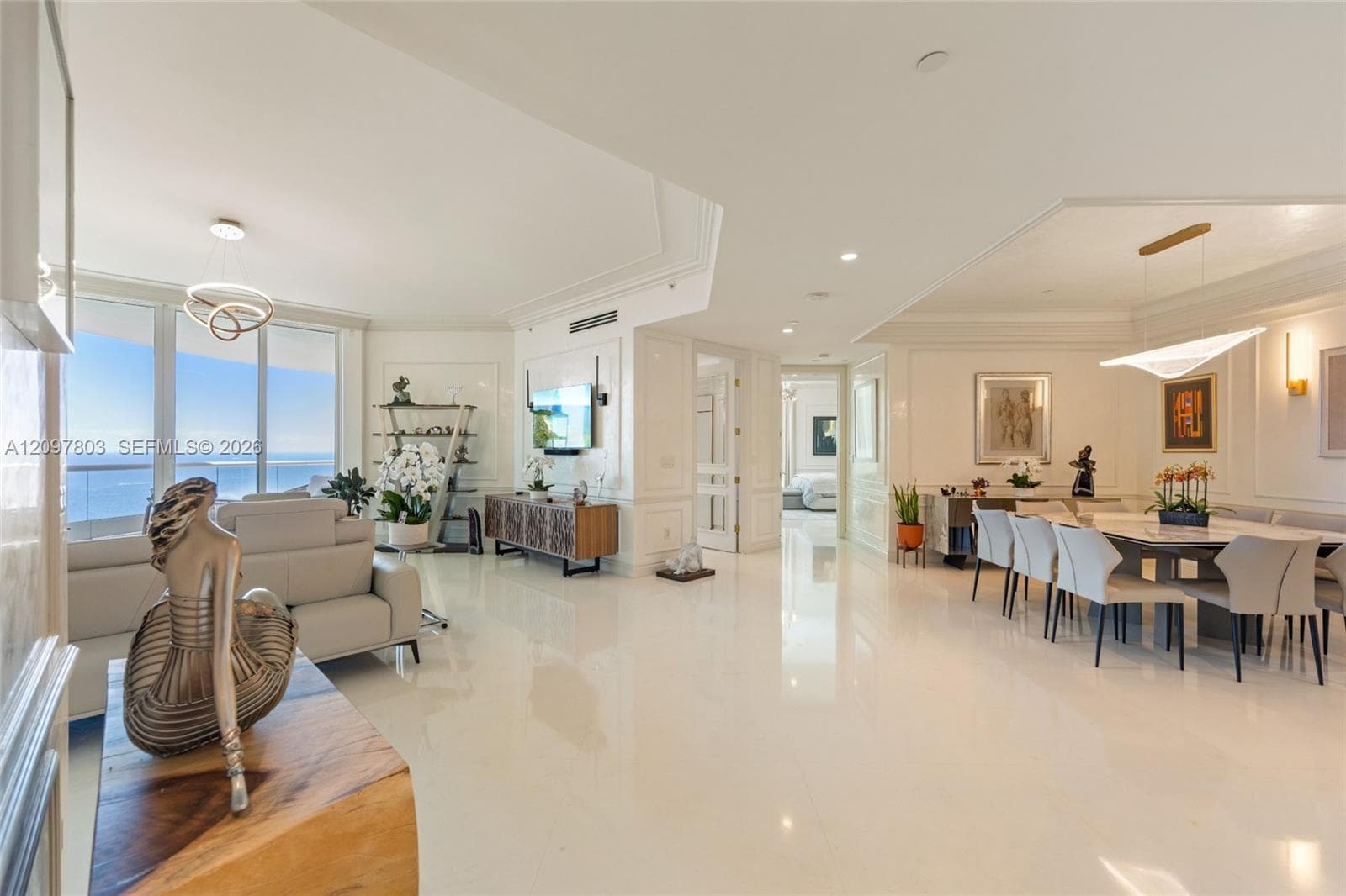 Open-concept living area featuring polished light-toned flooring, intricate tray ceilings with recessed lighting, modern geometric pendant chandeliers, and floor-to-ceiling glass paneling