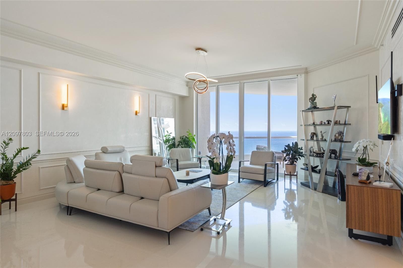 Living area featuring floor-to-ceiling windows, polished neutral-toned flooring, intricate wall molding, modern pendant lighting, and wall-mounted sconces