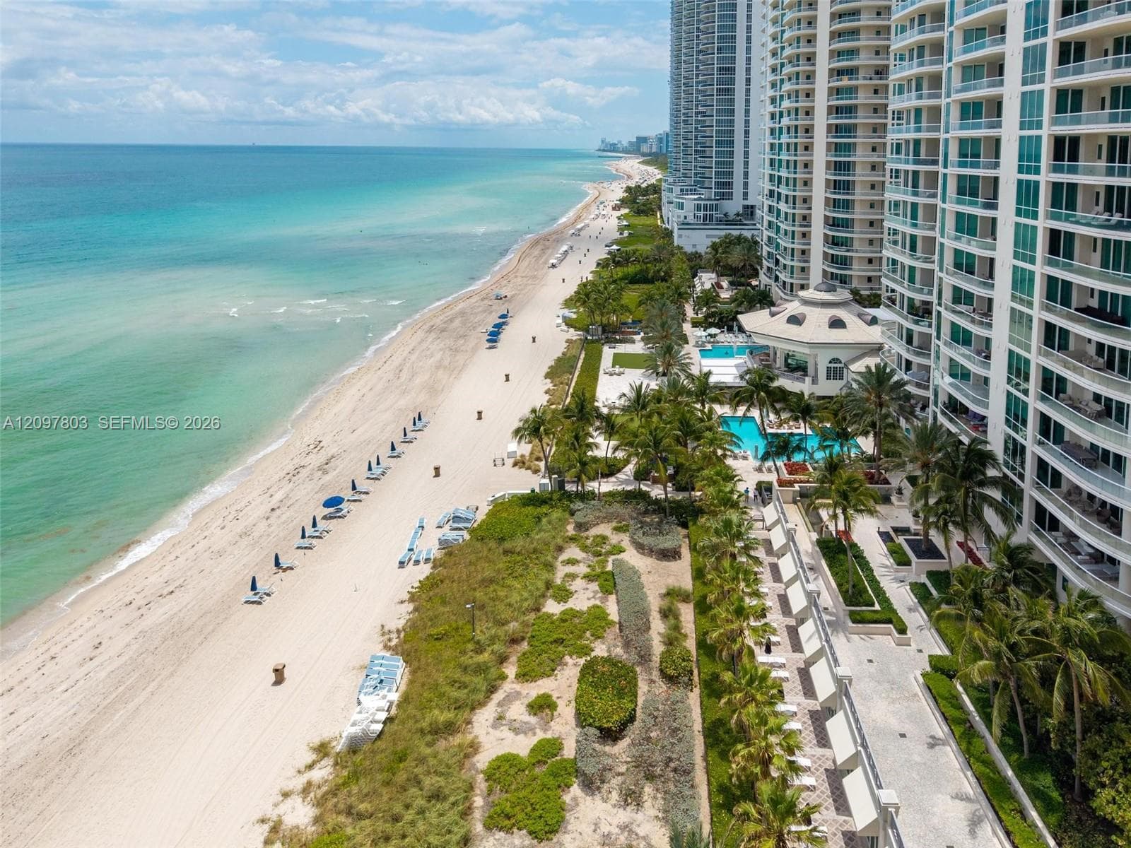 High-rise coastal property featuring direct beach access, outdoor swimming pools, stone-paved walkways, and established palm tree landscaping