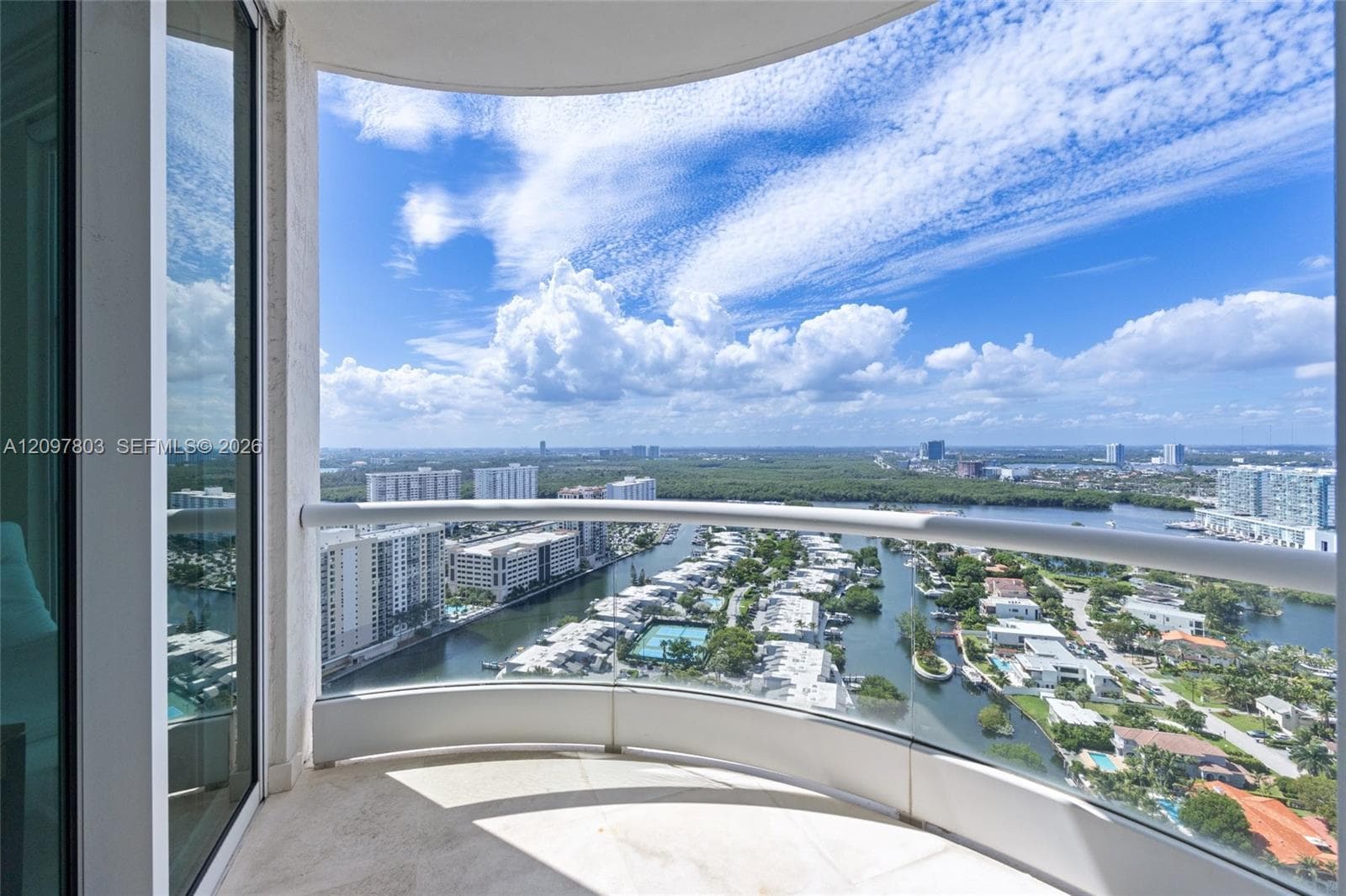 Curved private balcony with a glass and metal railing, stone flooring, and expansive water and urban scenery