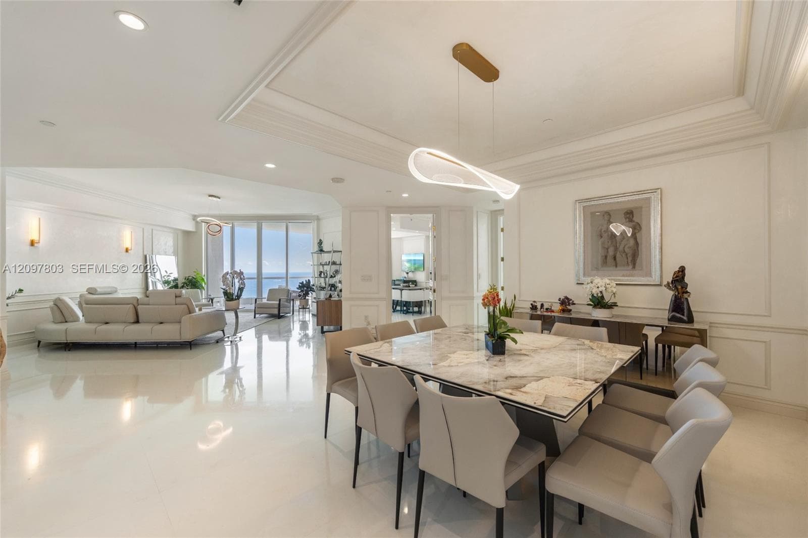 Open-concept interior featuring large-format polished tile flooring, tray ceilings with crown molding, custom wall paneling, and contemporary sculptural light fixtures