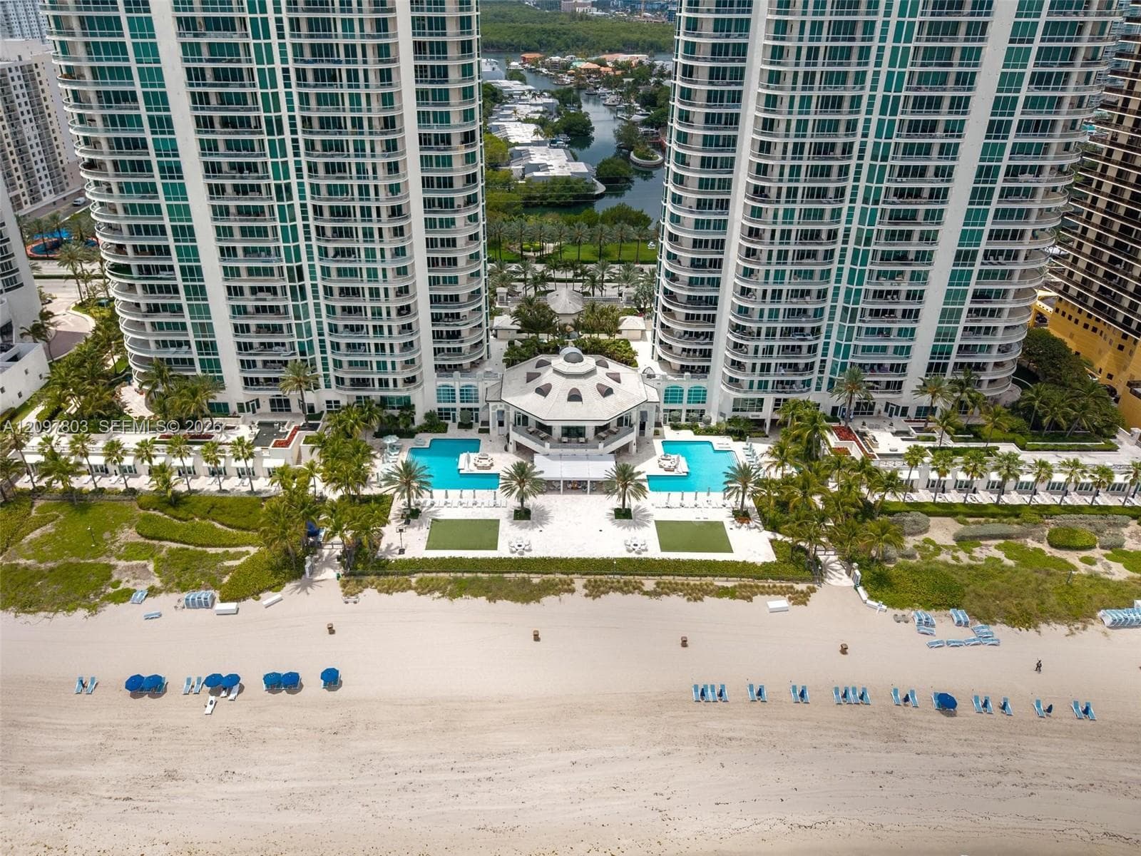 High-rise residential towers featuring beachfront access, twin swimming pools, a central pavilion, and manicured grounds